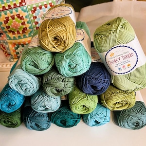 May include: A collection of 100% cotton yarn balls in various shades of green and blue, stacked in a pyramid shape. Each ball features a white paper label with the text 100% Cotton Yarn Chunky Thread by Lori Holt of Bee in my Bonnet.