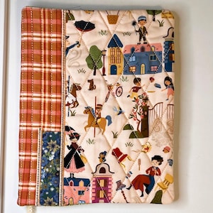 May include: A quilted fabric book cover featuring a cream-colored panel with a whimsical print of people, houses, and trees, accented by a vertical strip of orange and white plaid fabric and a smaller section of blue floral patterned fabric.