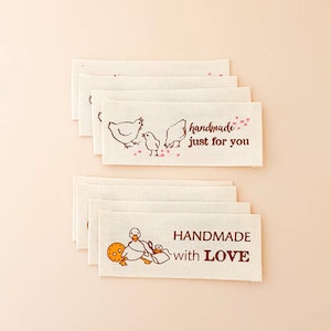 May include: A stack of rectangular off-white fabric labels featuring brown ink illustrations and text. The top set shows chickens and the text "handmade just for you" with pink accents. The bottom set shows a duck and "HANDMADE with LOVE".