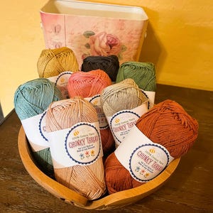 May include: A wooden bowl filled with colorful balls of chunky cotton yarn. The yarn is in various colors, including teal, peach, and rust. Each ball has a label that reads "CHUNKY THREAD" and "100% Cotton Yarn". A decorative floral container is in the background.