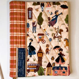 May include: A quilted fabric book cover featuring a cream-colored panel with a repeating pattern of London-themed illustrations, including a red telephone box, Big Ben, and a double-decker bus, alongside a vertical orange plaid panel and blue text.