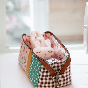 May include: A small, open zippered fabric box with a brown zipper and a brown and white gingham pattern. The box contains a stuffed bunny toy. The box has a patchwork design with floral and polka dot patterns.