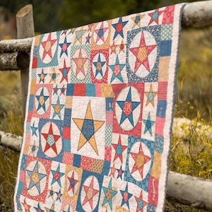 May include: A colorful quilt with a star pattern. The quilt features various star designs in red, blue, yellow, and white, set against a patchwork background. The quilt is draped over a wooden fence.