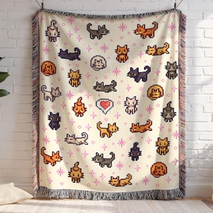 May include: A cream-colored woven throw blanket featuring a repeating pattern of pixelated cats in various poses and colors, including orange, black, and gray. Pink star accents and a heart graphic are also present. The blanket has a fringed edge.