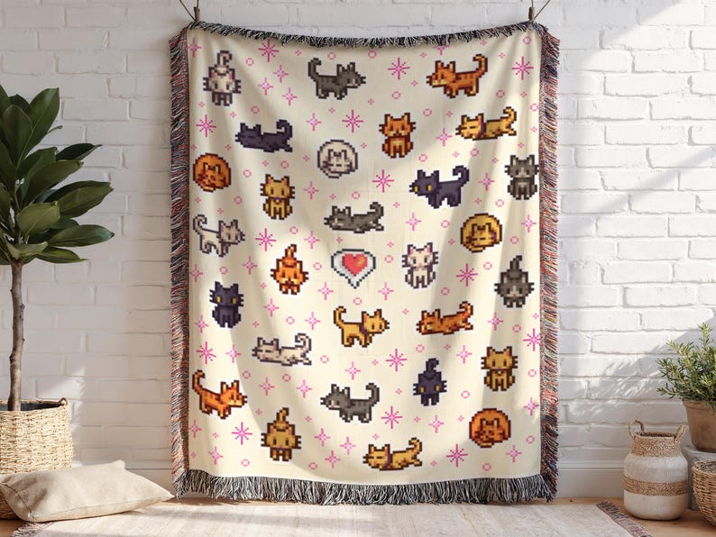 May include: A cream-colored woven throw blanket featuring a repeating pattern of pixelated cats in various poses and colors, including orange, black, and gray. Pink star accents and a heart graphic are also present. The blanket has a fringed edge.