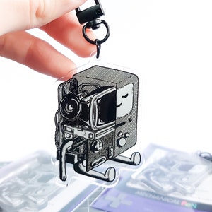 Könnte beinhalten: Ein transparenter Acryl-Schlüsselanhänger mit einer Schwarzweiß-Illustration einer Retro-Videospielkonsole. Die Konsole besteht aus verschiedenen Teilen, darunter ein Fernseher, eine Kamera und eine Spielkassette. Der Schlüsselanhänger hat einen schwarzen Metall-Drehverschluss.