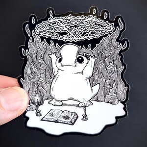 May include: A black and white illustration of a cartoon platypus performing a ritual with a pentagram, candles, and a book with a pentagram on the cover. The platypus is surrounded by flames.