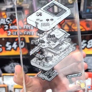 Puede incluir: Panel de acrílico transparente con una ilustración detallada en vista explosionada de una consola de juegos portátil clásica. El diseño incluye componentes internos, baterías y el texto "MECHANICAL PEN". El panel está sujeto con tornillos dorados.