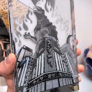 Puede incluir: Bloque acrílico transparente con una ilustración en blanco y negro de Godzilla destruyendo una ciudad. La obra de arte incluye texto japonés y llamas. El bloque está sujeto con tornillos dorados.