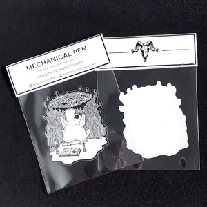 Puede incluir: Una pegatina en blanco y negro con una criatura de dibujos animados con una corona de llamas y un libro. La pegatina est&aacute; empaquetada en una bolsa de pl&aacute;stico transparente con una etiqueta blanca que dice "MECHANICAL PEN Imagine | Create | Inspire @MechanicalPen_ mechanicalpen.etsy.com".