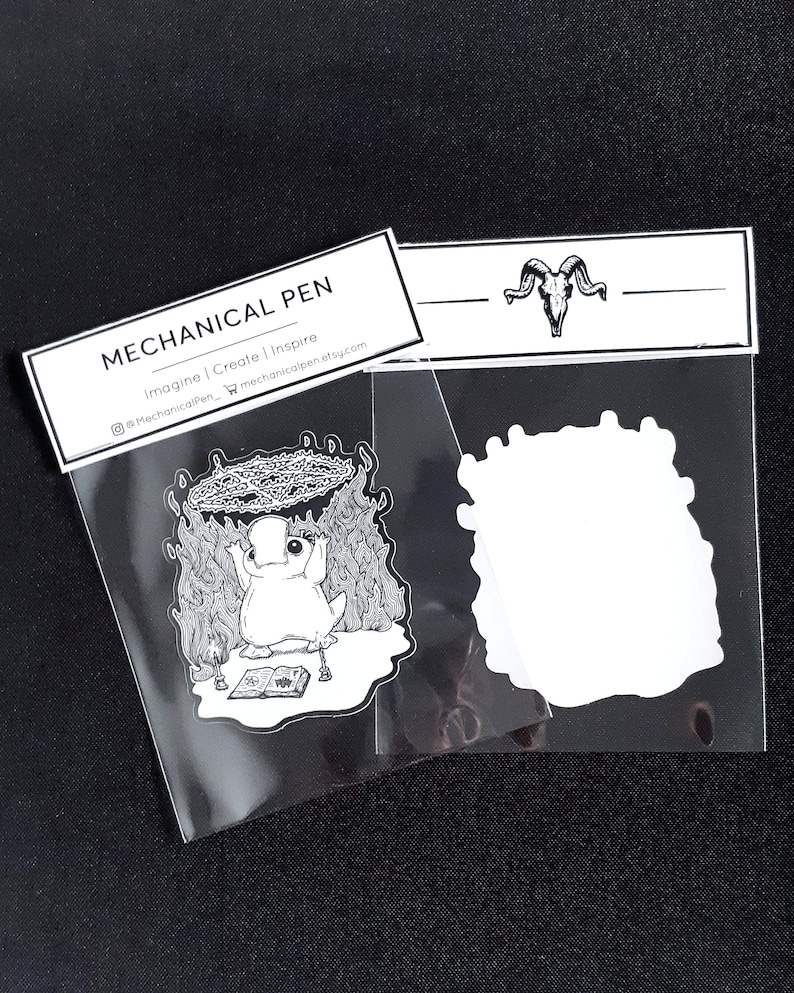 Puede incluir: Una pegatina en blanco y negro con una criatura de dibujos animados con una corona de llamas y un libro. La pegatina est&aacute; empaquetada en una bolsa de pl&aacute;stico transparente con una etiqueta blanca que dice "MECHANICAL PEN Imagine | Create | Inspire @MechanicalPen_ mechanicalpen.etsy.com".