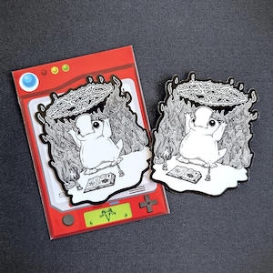 May include: A black and white enamel pin featuring a cartoon character with a crown of flames and a book in front of it. The pin is on a red and white background.