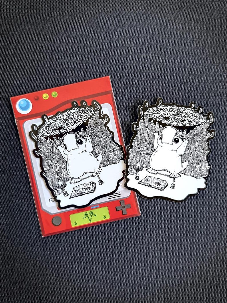May include: A black and white enamel pin featuring a cartoon character with a crown of flames and a book in front of it. The pin is on a red and white background.