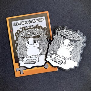 May include: A black and white sticker featuring a cartoon character with a circle of flames above its head. The sticker is on a yellow and orange background with the text "Premium Matte Vinyl Mechanical Pen".