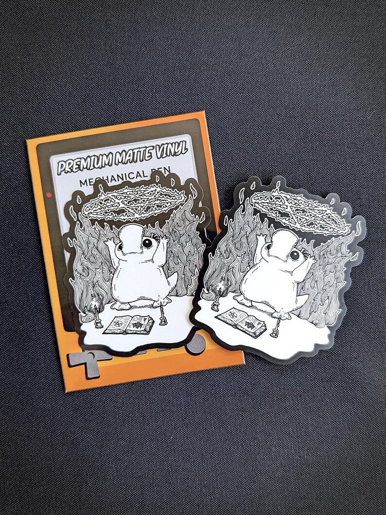 May include: A black and white sticker featuring a cartoon character with a circle of flames above its head. The sticker is on a yellow and orange background with the text "Premium Matte Vinyl Mechanical Pen".