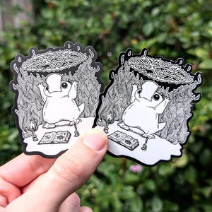 May include: Two black and white stickers featuring a cartoon frog wearing a crown and sitting on a book with flames and a pentagram around it. The stickers are on a green background.