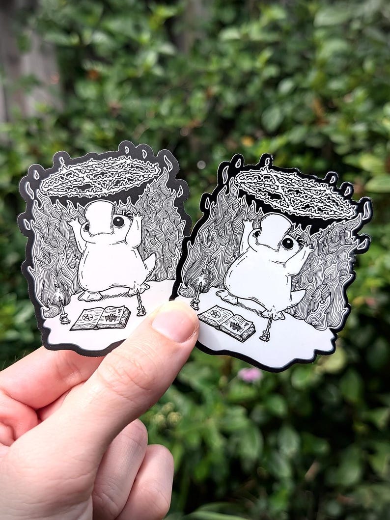 May include: Two black and white stickers featuring a cartoon frog wearing a crown and sitting on a book with flames and a pentagram around it. The stickers are on a green background.