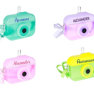 May include: Four colorful camera-shaped cases, each with a different color scheme: teal, lavender, pink, and yellow-green. Each case has a lens, a clear tube, and the name "Alexander" printed on top. A ribbon or strap is attached to each case.