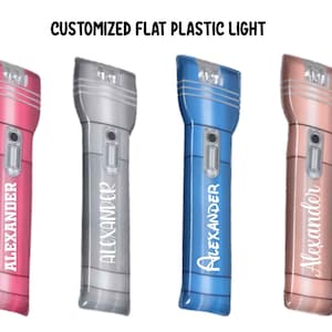 May include: Four flat plastic flashlights in pink, silver, blue, and rose gold. Each flashlight has the name "Alexander" printed vertically. The flashlights have a rectangular shape with a rounded top and a small button.
