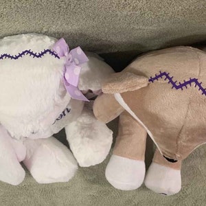 May include: Two plush toys, one white with purple and white polka dot bows and a purple zig-zag stitch, and one brown with a purple zig-zag stitch.