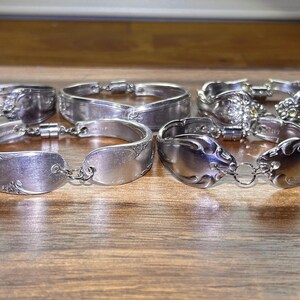 May include: A set of eight silver spoon bracelets with intricate designs. The bracelets are linked together with a chain.