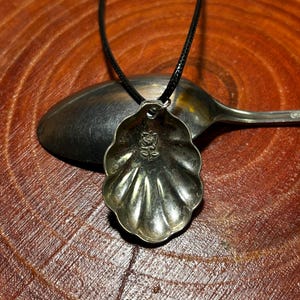 May include: A silver pendant necklace with a shell-shaped pendant and a floral design. The pendant is suspended from a black cord. The necklace is shown on a wooden surface with a spoon.