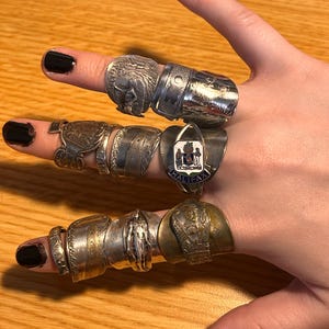 May include: A hand wearing a variety of silver and gold rings. The rings feature different designs, including a crest with the text "HALIFAX" and a design of a person.
