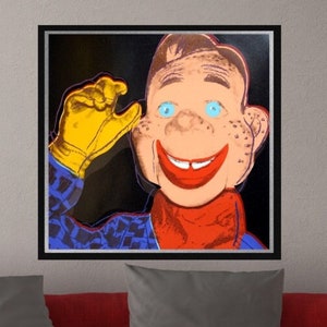May include: Pop art print of a smiling cartoon character with blue eyes, freckles, and a red mouth. The character wears a red scarf, a blue patterned shirt, and a yellow glove. The artwork is framed in black and hangs above a red sofa.
