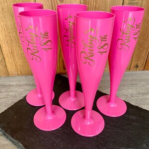 May include: Five pink plastic champagne flutes with gold lettering that reads "Ruby's 18th".