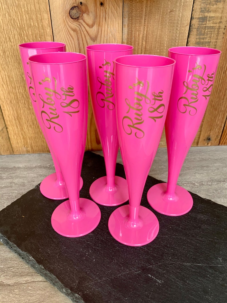 May include: Five pink plastic champagne flutes with gold lettering that reads "Ruby's 18th".