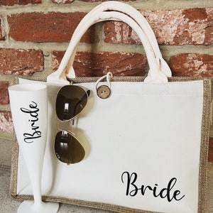 May include: A white canvas tote bag with brown trim and the word "Bride" printed in black. The bag has a white rope handle and a brown button closure. A pair of sunglasses and a white champagne flute are in front of the bag.