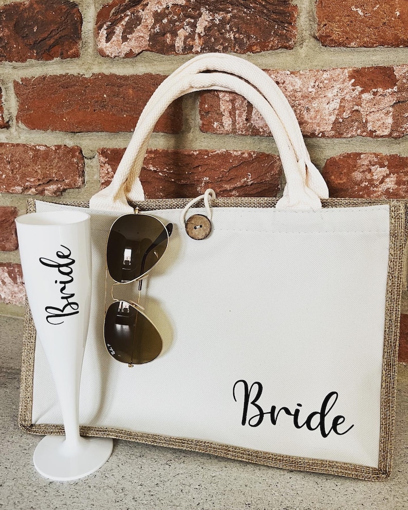 May include: A white canvas tote bag with brown trim and the word "Bride" printed in black. The bag has a white rope handle and a brown button closure. A pair of sunglasses and a white champagne flute are in front of the bag.