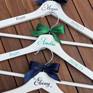 May include: Three white wooden hangers with the words "Prom" and the dates "29/06/23" and "06/07/23" printed on them. Each hanger has a different colored ribbon bow: navy blue, green, and navy blue. The hangers are personalized with the names "Megan", "Amelia", and "Ebony".