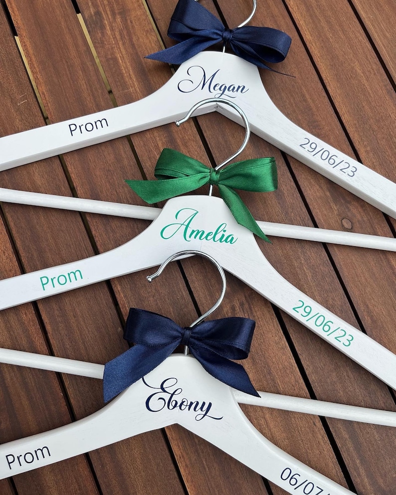 May include: Three white wooden hangers with the words "Prom" and the dates "29/06/23" and "06/07/23" printed on them. Each hanger has a different colored ribbon bow: navy blue, green, and navy blue. The hangers are personalized with the names "Megan", "Amelia", and "Ebony".
