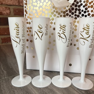 May include: Four white plastic champagne flutes with gold lettering. The glasses are personalised with the names Louise, Zoe, Beth, and Katie, along with the titles Maid of Honour, Bride, and Bridesmaid printed in a script and sans-serif font.