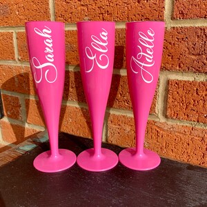 May include: Three pink plastic champagne flutes with white vinyl lettering. The flutes are labeled "Sarah", "Ella", and "Adelle".