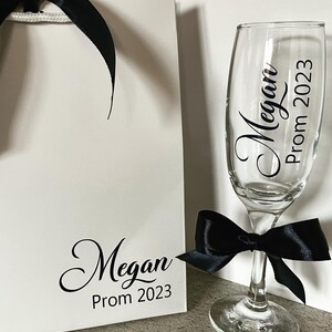 May include: A white gift bag with a black ribbon and the text "Megan Prom 2023". Next to the bag is a champagne flute with a black ribbon and the text "Megan Prom 2023".