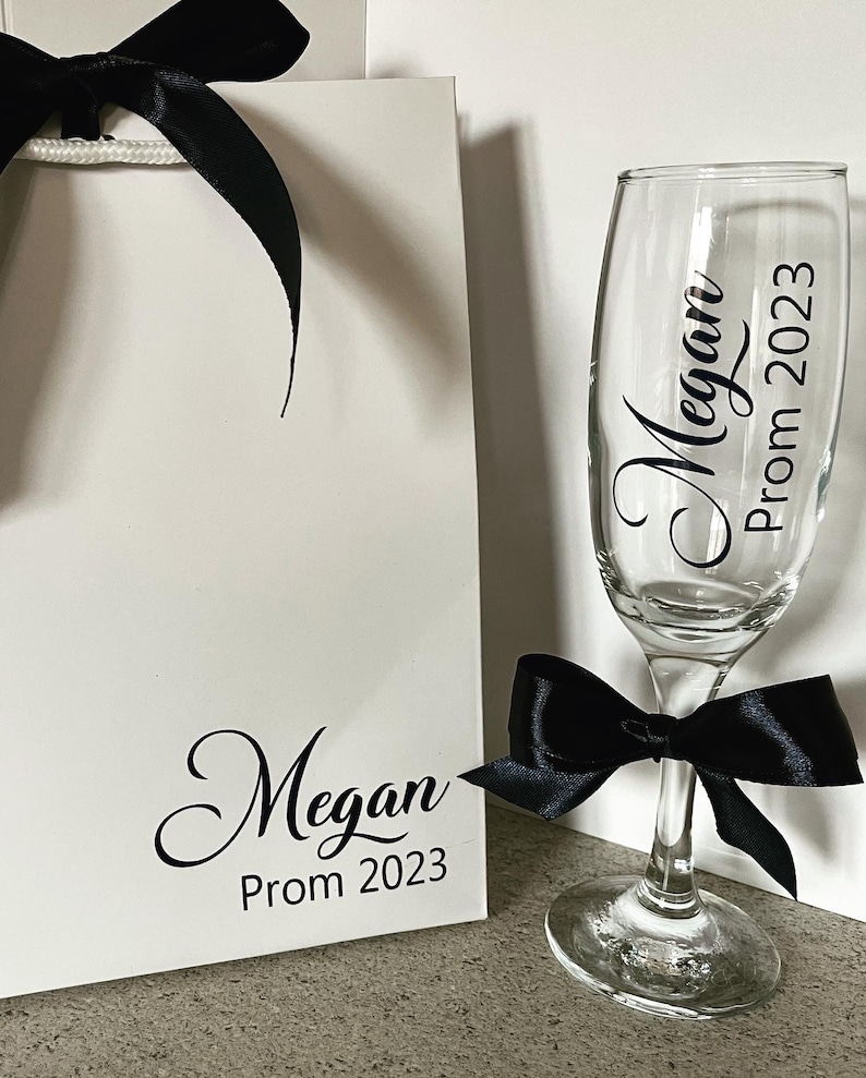 May include: A white gift bag with a black ribbon and the text "Megan Prom 2023". Next to the bag is a champagne flute with a black ribbon and the text "Megan Prom 2023".