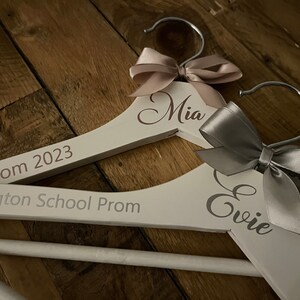 May include: Two white wooden hangers with personalized names "Mia" and "Evie" and the text "Prom 2023" and "Eckington School Prom". Each hanger is decorated with a satin ribbon bow.