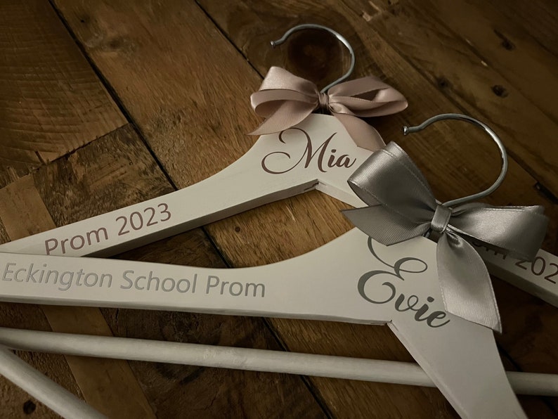 May include: Two white wooden hangers with personalized names "Mia" and "Evie" and the text "Prom 2023" and "Eckington School Prom". Each hanger is decorated with a satin ribbon bow.