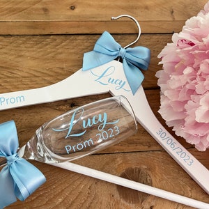 May include: White wooden hanger with a blue bow and the text "Lucy Prom 2023" and "30/06/2023". A champagne flute with a blue bow and the text "Lucy Prom 2023" is resting on the hanger.