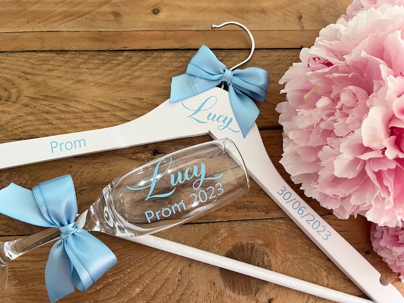 May include: White wooden hanger with a blue bow and the text "Lucy Prom 2023" and "30/06/2023". A champagne flute with a blue bow and the text "Lucy Prom 2023" is resting on the hanger.