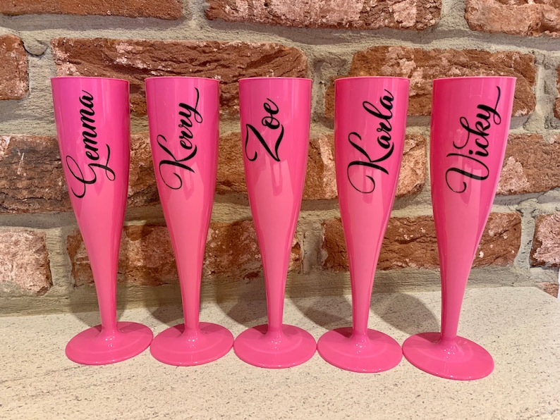 May include: Five pink plastic champagne flutes with black lettering. The flutes are personalized with the names "Gemma", "Kerry", "Zoe", "Karla", and "Vicky".