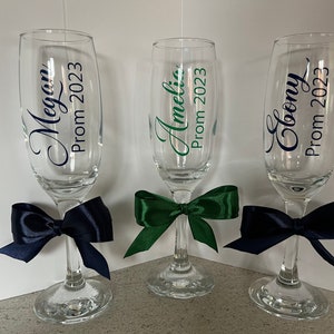 May include: Three champagne flutes with personalized labels for a prom celebration. The flutes are decorated with navy blue and green bows. The labels read "Megan Prom 2023", "Amelia Prom 2023", and "Cloony Prom 2023".