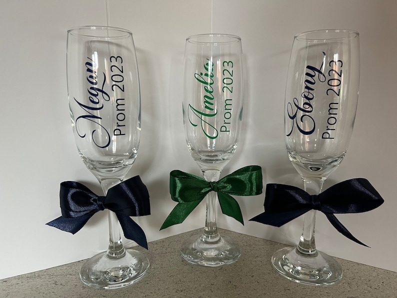 May include: Three champagne flutes with personalized labels for a prom celebration. The flutes are decorated with navy blue and green bows. The labels read "Megan Prom 2023", "Amelia Prom 2023", and "Cloony Prom 2023".
