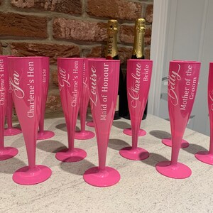 May include: Pink plastic champagne flutes with white lettering. The flutes are personalized with names and titles such as "Bride", "Maid of Honor", and "Mother of the Groom".