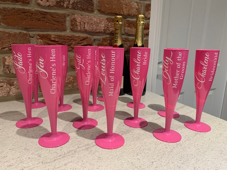 May include: Pink plastic champagne flutes with white lettering. The flutes are personalized with names and titles such as "Bride", "Maid of Honor", and "Mother of the Groom".