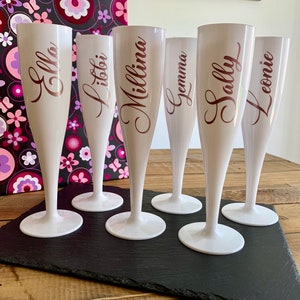 May include: Six white plastic champagne flutes with gold lettering. The flutes are arranged in a row on a black slate surface. The names on the flutes are Ella,  Litoti, Millina, Gemma, Sally, and Leonie.