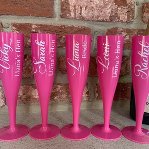 May include: Five pink plastic champagne flutes with white lettering. The flutes are personalized with names and the phrase "Liana's Hen".