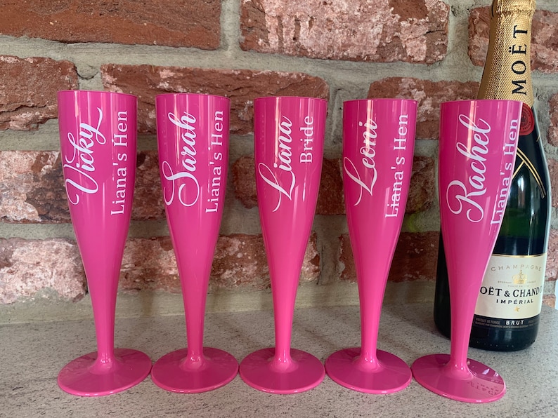May include: Five pink plastic champagne flutes with white lettering. The flutes are personalized with names and the phrase "Liana's Hen".